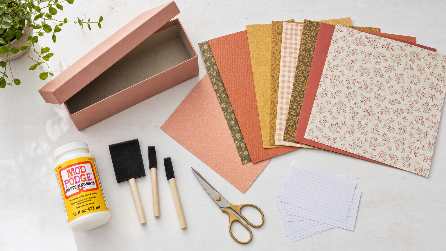 Flat lay of decoupage supplies: Mod Podge bottle, foam brushes, gold scissors, patterned scrapbook papers in coral and mustard, pink box, index cards.