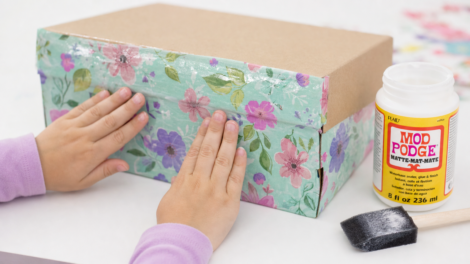 Child's hands smoothing mint floral decoupage paper onto a cardboard shoebox, with Mod Podge matte and a foam brush nearby.